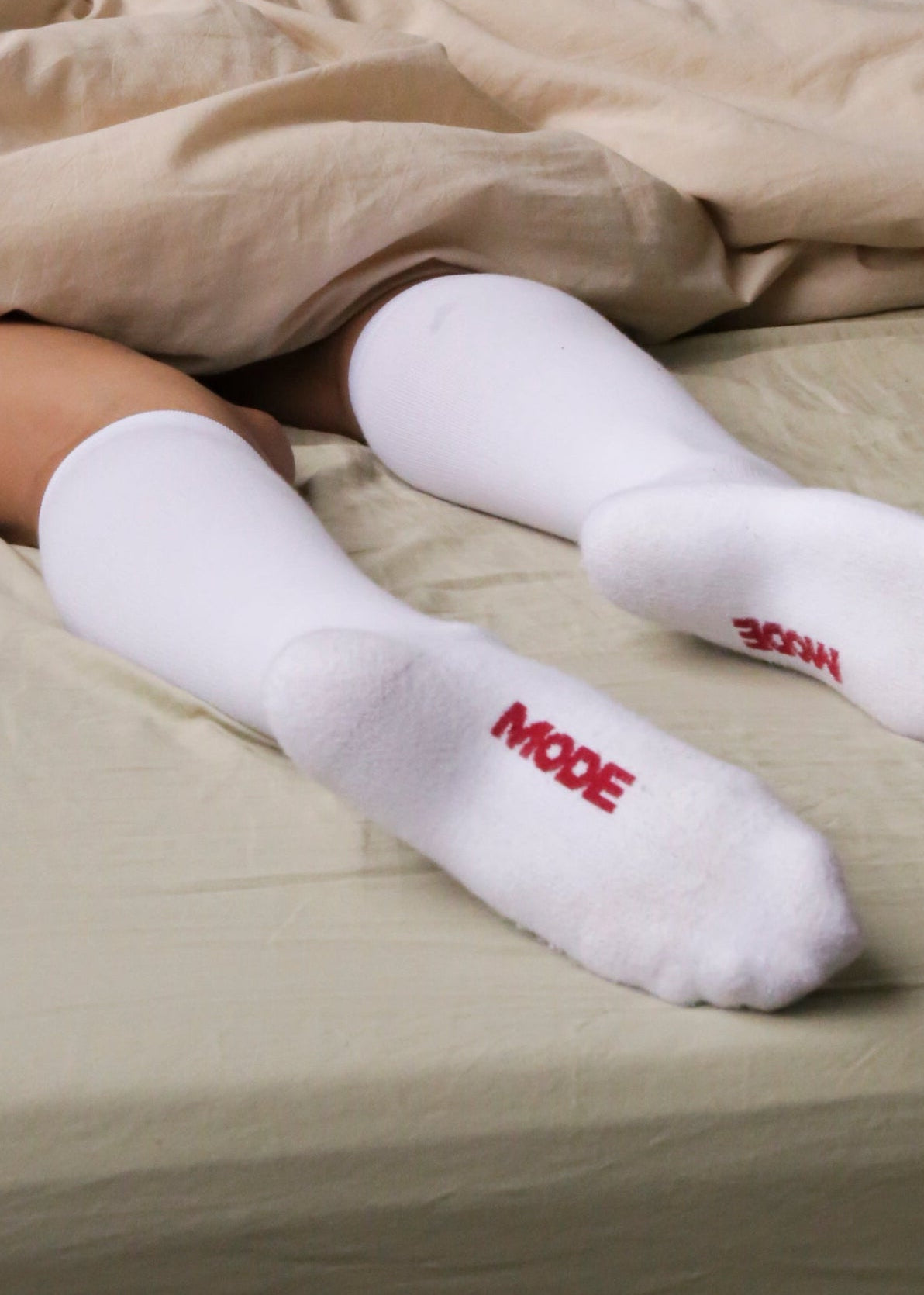 Person lying in bed wearing white socks with red text, covered by beige sheets.