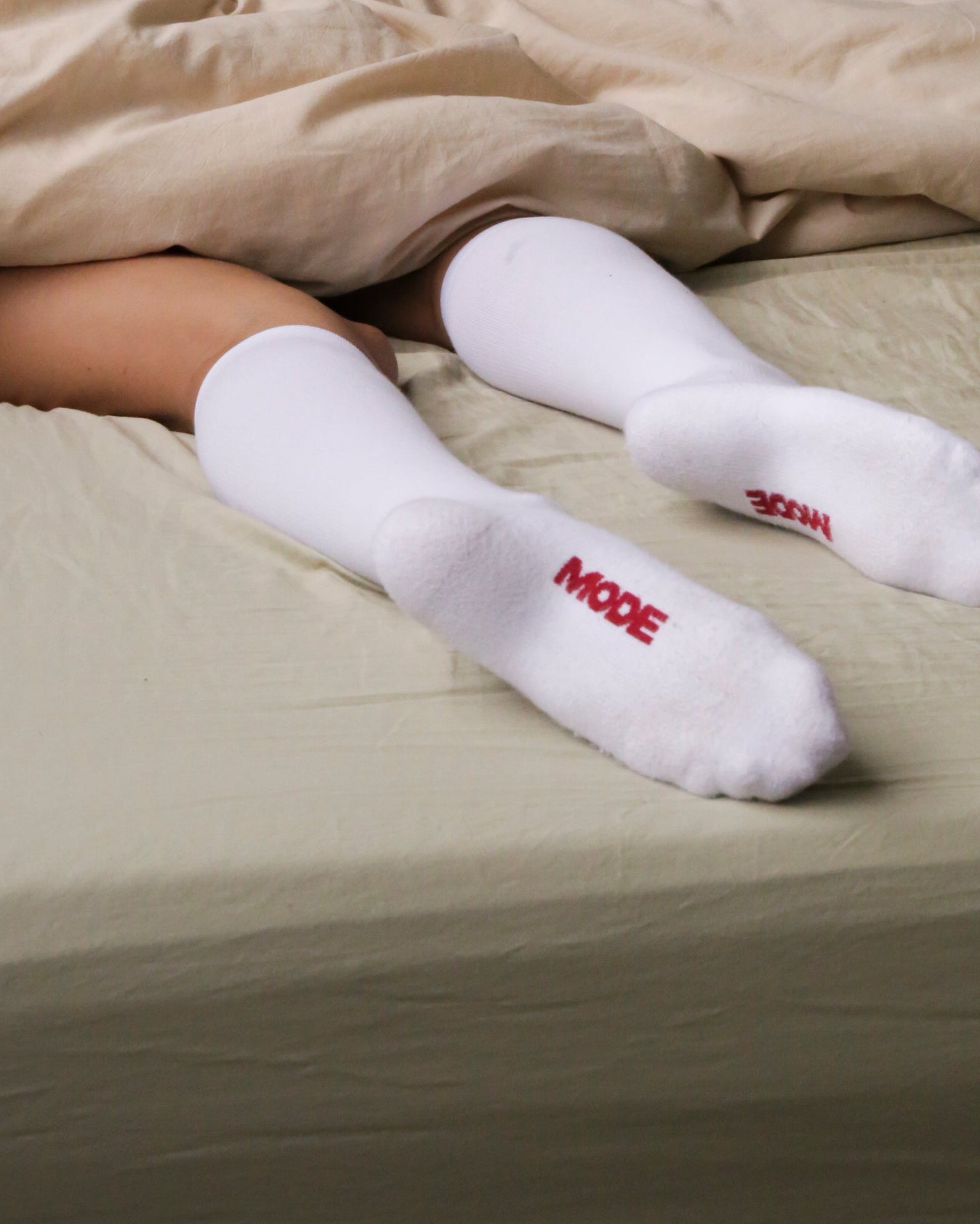Person lying in bed wearing white socks with red text, covered by beige sheets.