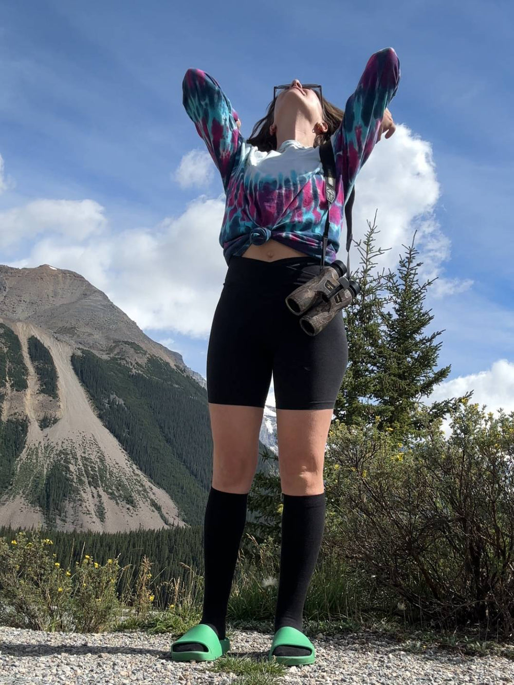 Person in colorful tie-dye hoodie and black shorts standing outdoors with mountains and sky in the background