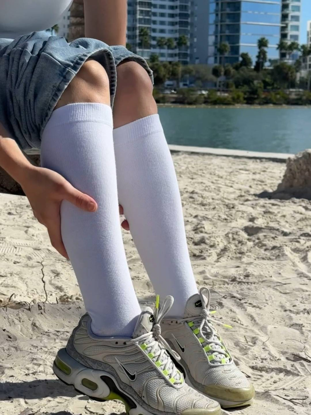 Person wearing knee-high socks and sneakers on a sandy surface with a lake and buildings in the background
